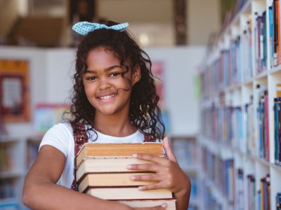 portrait-of-smiling-african-american-schoolgirl-carrying-stack-of-books-in-school-library.jpg